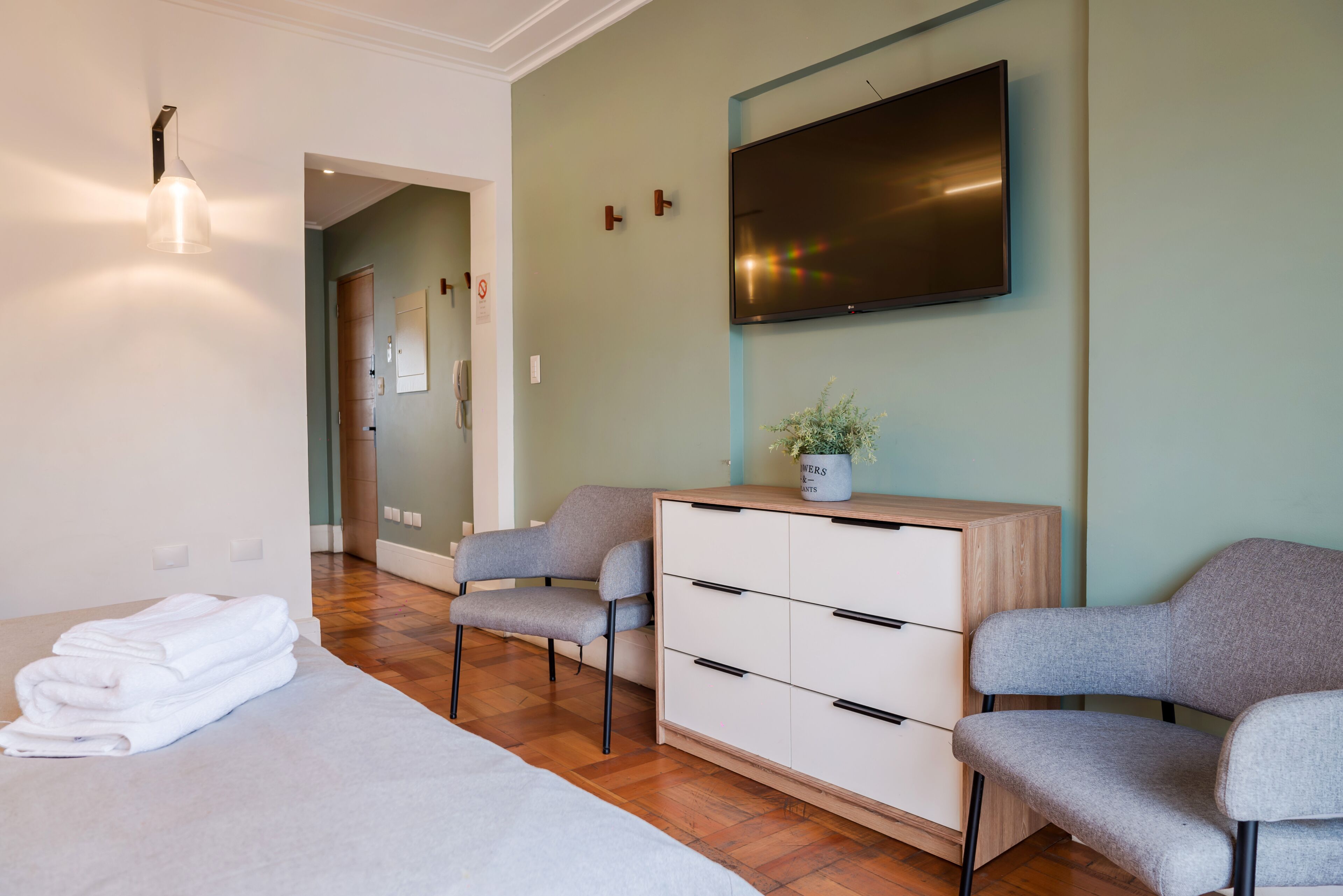 Quarto de hotel moderno com uma cama, duas poltronas cinzas, uma cômoda de madeira, uma TV de tela plana na parede e um vaso de plantas na cômoda.