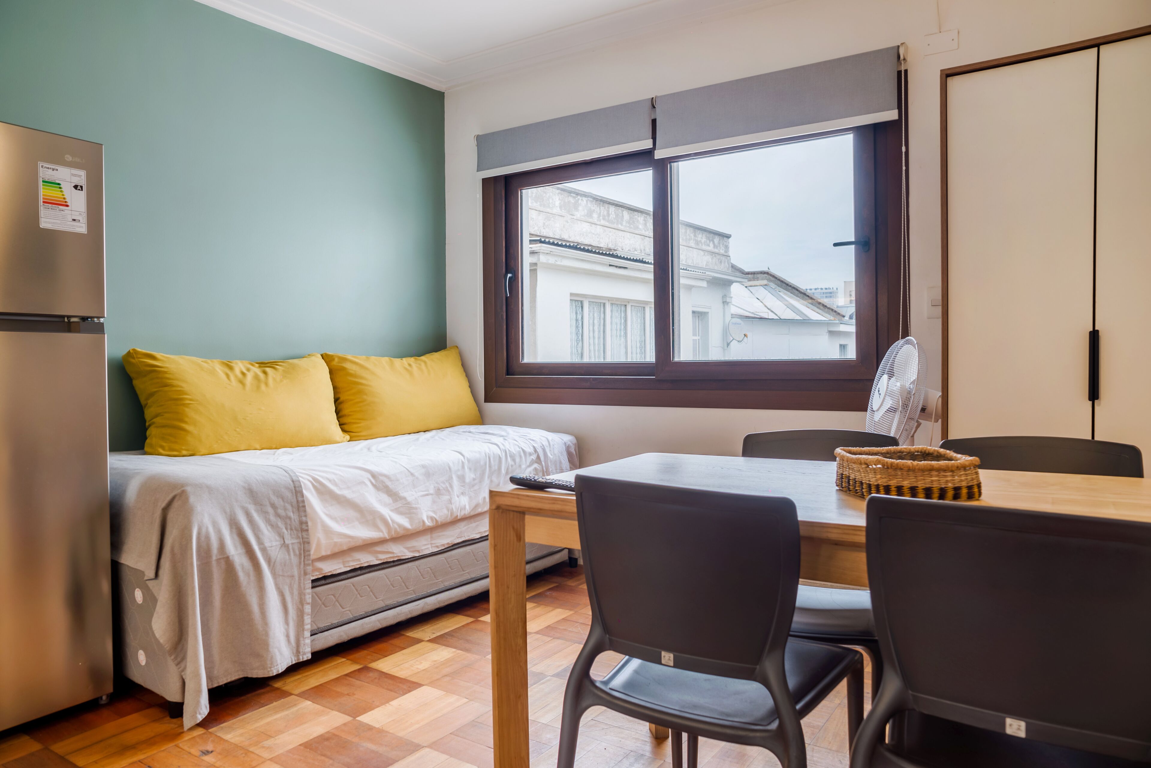 Ein kleines Zimmer mit einem Bett, einem Tisch, zwei Stühlen, einem Fenster und einem Kühlschrank.