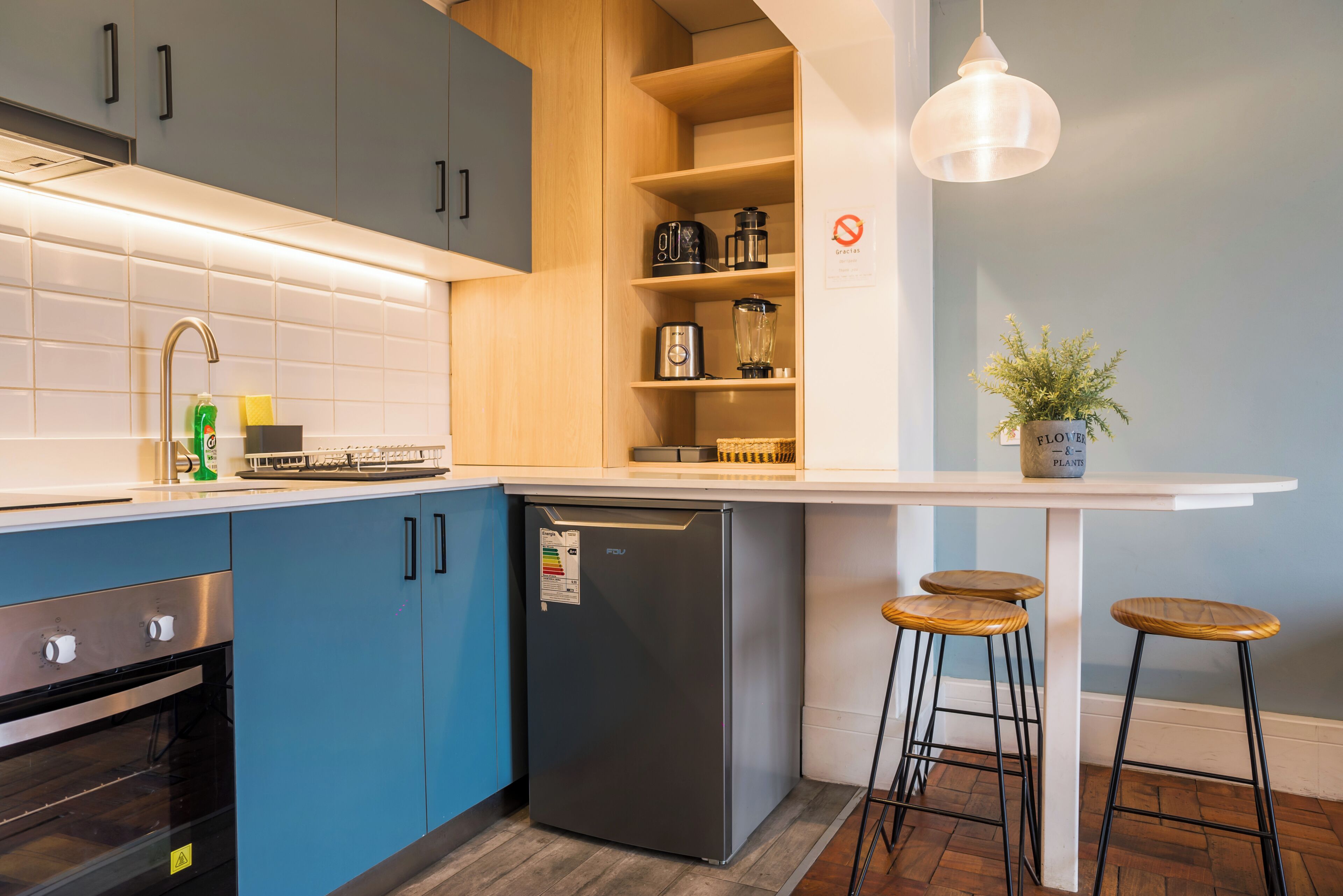 A modern kitchen with blue cabinets, a built-in oven, a refrigerator, and a bar area with stools.