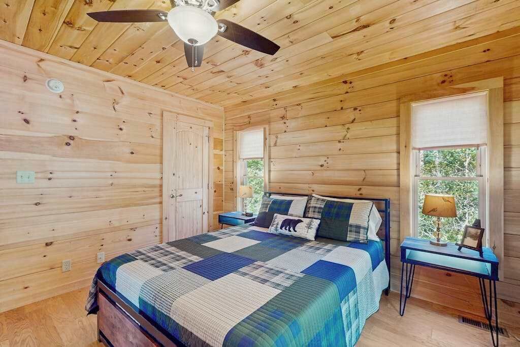 A wooden cabin bedroom with a bed, a nightstand, a ceiling fan, and a window with a view of trees.