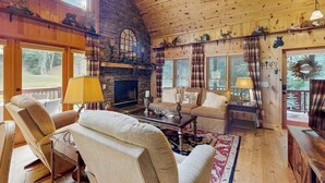 Interior acogedor de una cabaña con chimenea de piedra, techo de madera y ventanas amplias.