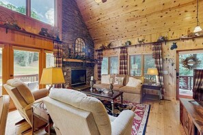 A cozy cabin interior with a stone fireplace, wooden walls, a large window, and a comfortable seating area.