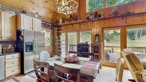 Interior de una cabaña de madera con comedor, cocina y grandes ventanales que ofrecen vistas al exterior.