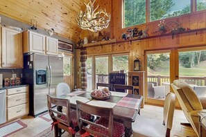 Interior de una cabaña de madera con comedor, cocina y ventanas amplias que ofrecen vistas al exterior.