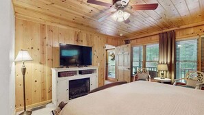 Una habitación con paneles de madera, una cama, un televisor sobre un mueble, un ventilador de techo y una ventana con persianas.