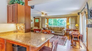 A kitchen with granite countertops, wooden cabinets, a dining area with a table and chairs, a sofa, and a large window with a view of trees. - The Rock House at Big Bear Estates (Waynesville)