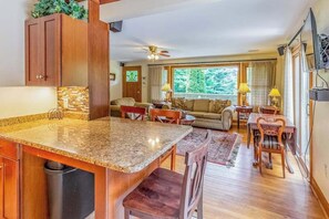 Una cocina con encimeras de granito, armarios de madera, un comedor con mesa y sillas, un sofá y un amplio ventanal con vista a árboles.
