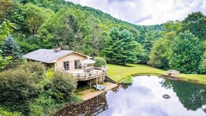 A house with a deck overlooking a pond, surrounded by lush greenery and hills. - The Rock House at Big Bear Estates (Waynesville)