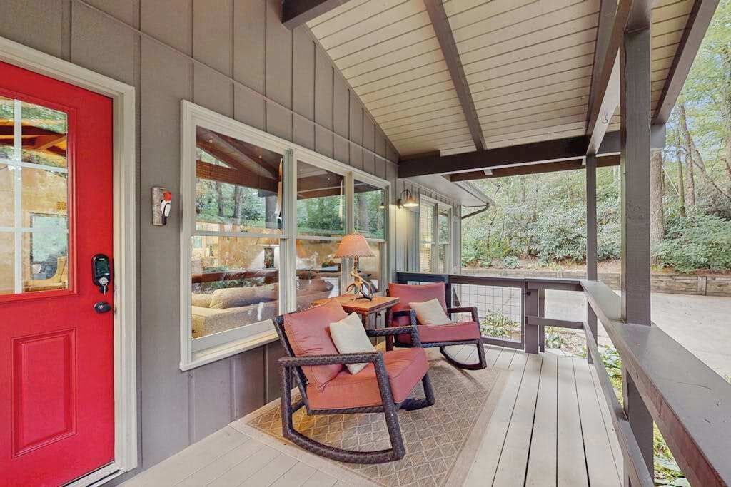 A covered porch with a red door, two rocking chairs, a table, and a lamp.