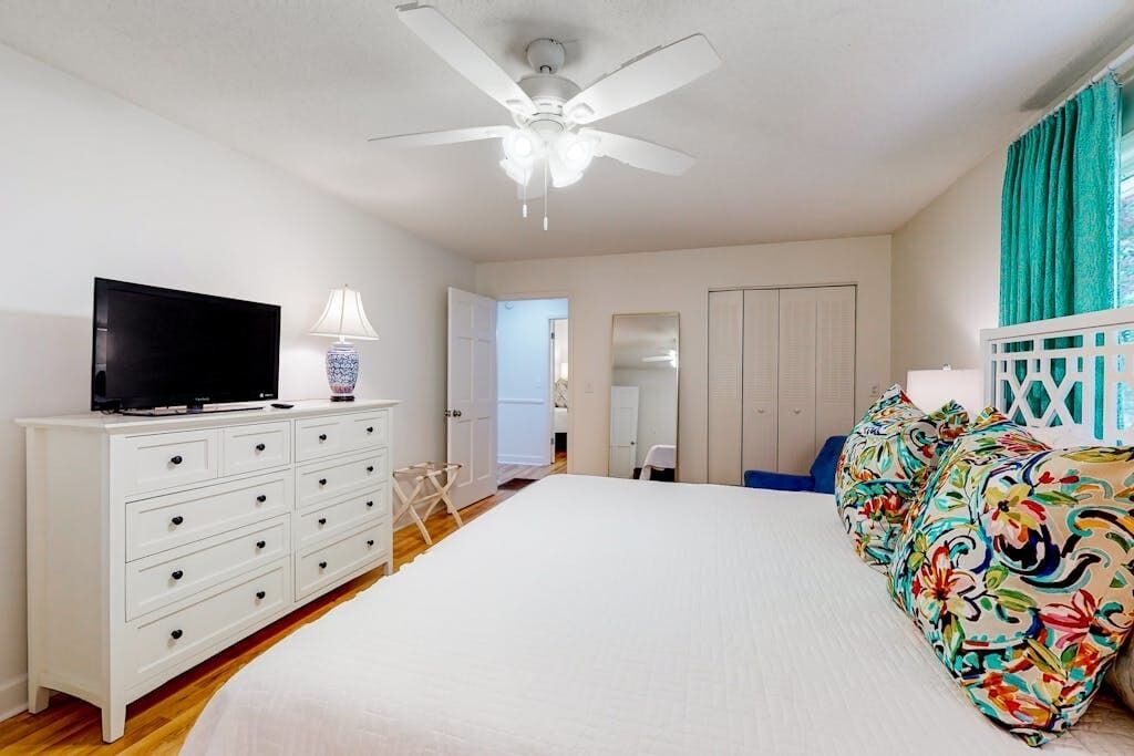 A bedroom with a bed, a TV on a dresser, a ceiling fan, and a door leading to another room.
