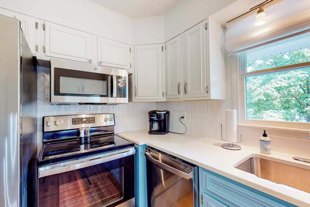 A kitchen with white cabinets, a microwave, an oven, a dishwasher, and a window with a view of trees.