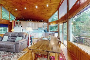 A cozy cabin interior with a wooden ceiling, a dining area, a sofa, and a kitchen with large windows.