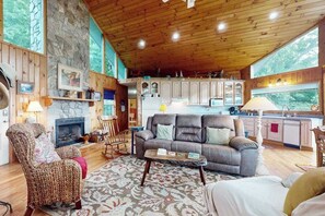 A cozy cabin interior with a stone fireplace, wooden ceiling, and a kitchen area.