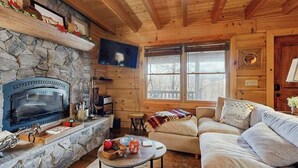 A cozy cabin interior with a stone fireplace, wooden walls, a television, a sofa, and a window. - Bear Hug (Sylva)