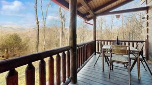 A wooden deck with a railing, a table with chairs, and a view of a forested landscape. - Bear Hug (Sylva)