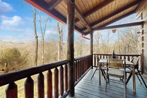 A wooden deck with a railing, a table with chairs, and a view of a forested landscape. - Bear Hug (Sylva)