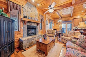 A log cabin interior with a stone fireplace, wooden furniture, and a ceiling fan. - Timberwolf (Tuckasegee)