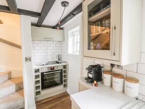 Cocina compacta con armarios blancos, horno integrado, cafetera y frascos de almacenamiento etiquetados con té, café y azúcar.