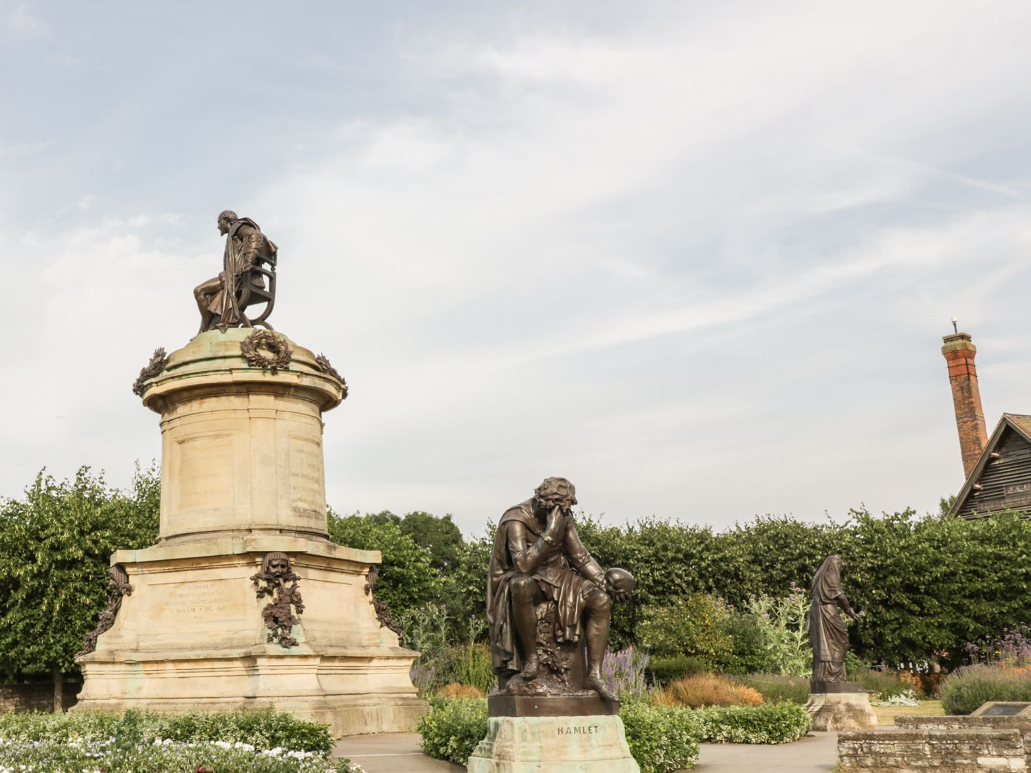 A statue of Hamlet on a pedestal, surrounded by greenery and a brick chimney in the background.