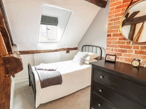 Un dormitorio con cama alta, una ventana pequeña, pared de ladrillo, viga de madera, cómoda y un reloj.