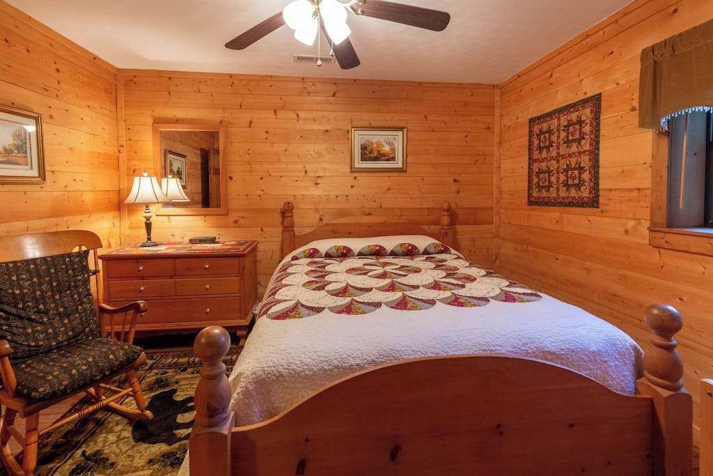 Un dormitorio de cabaña de madera con una cama, un tocador, una silla, un ventilador de techo y un edredón.