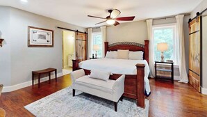 Quarto com cama grande, ventoinha no teto, um banco e janela com cortinas.