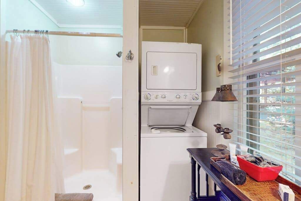 A laundry room with a washing machine and dryer, a small table with items, and a window with blinds.