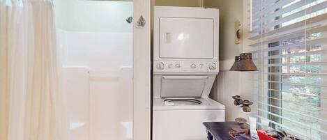 A laundry room with a washing machine and dryer, a small table with items, and a window with blinds.