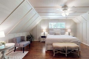 Ein Schlafzimmer mit schrägen Wänden, einem Bett mit weißer Bettdecke, einem Stuhl, einem Nachttisch mit Lampe und einem Fenster mit Jalousien.