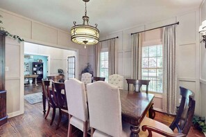 Ein Esszimmer mit Holztisch, gepolsterten Stühlen, einer Kronleuchter und großen Fenstern mit Vorhängen.