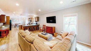 Un salón amplio con un sofá grande, un televisor montado en la pared y una cocina al fondo.