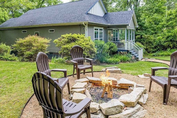 Ein Haus mit Feuerstelle, umgeben von Stühlen, einer überdachten Veranda und einem gepflegten Garten.