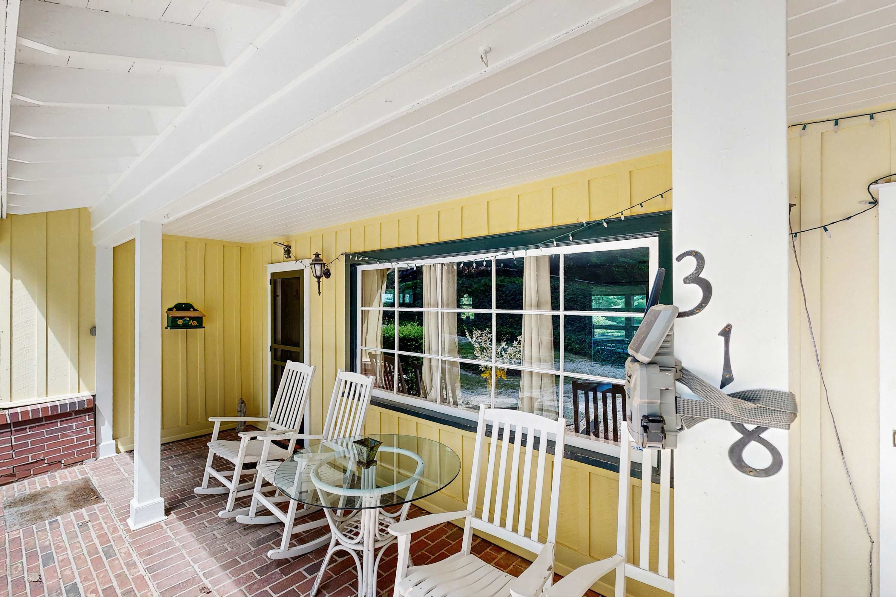 A covered patio with a glass table and white chairs, a brick wall, and a yellow exterior wall.