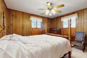 Ein Schlafzimmer mit Holzwandverkleidung, einem Bett mit weißer Bettwäsche, einem Holzstuhl, einem Schreibtisch und einem Deckenventilator.