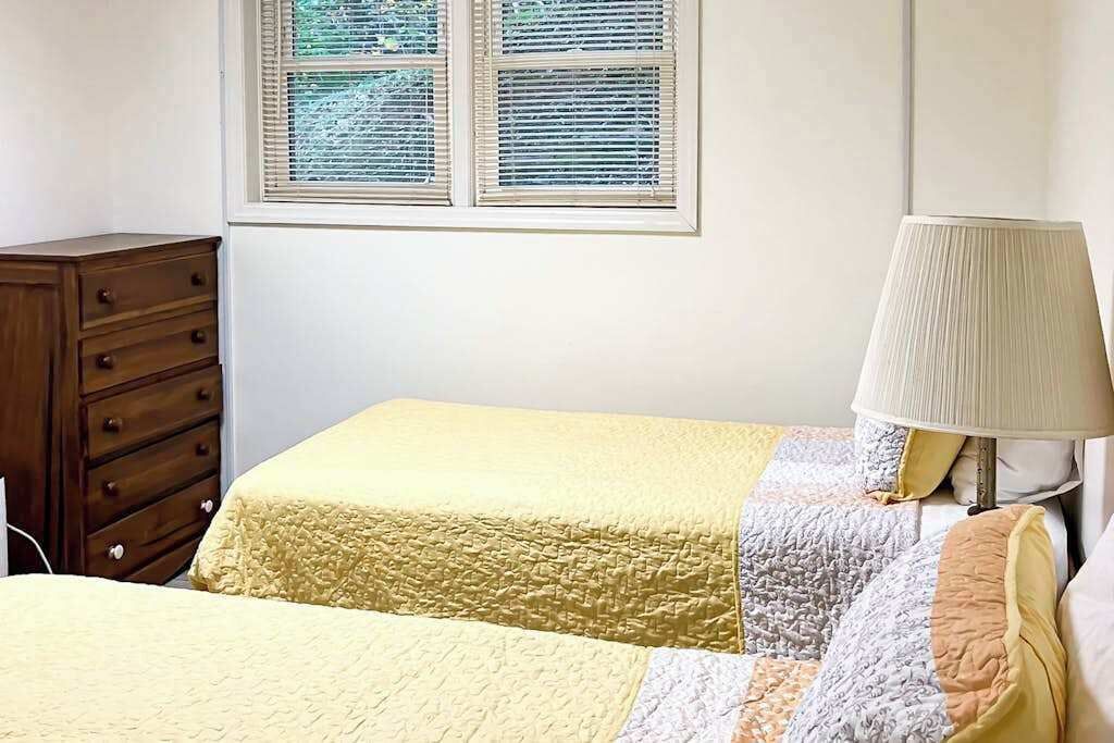 Une chambre à coucher comprenant un lit, une table de chevet, une lampe, une commode et une fenêtre avec des stores.