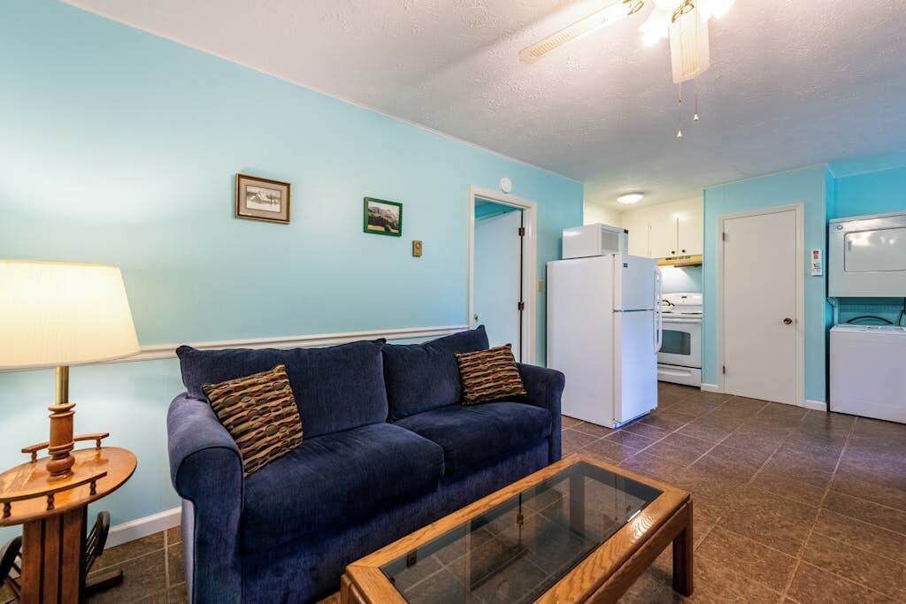 A living room with a blue couch, a glass coffee table, a lamp, and a kitchen area with a refrigerator and oven.