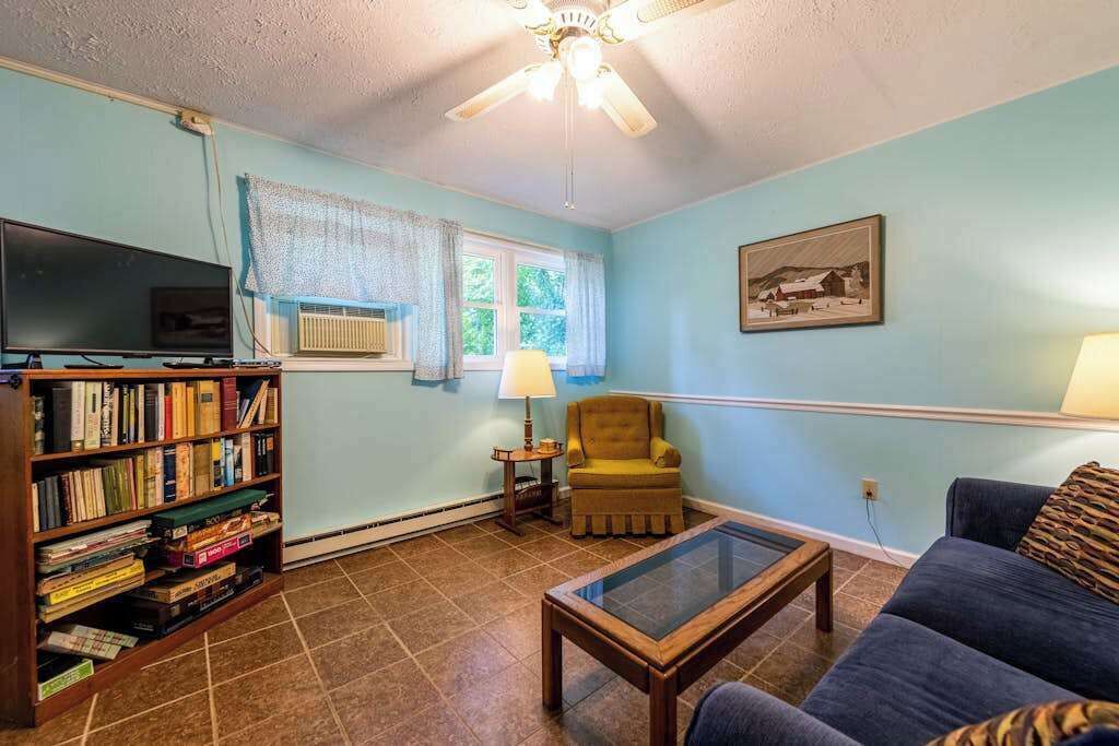 A living room with a bookshelf, a television, a ceiling fan, a window with curtains, a chair, a couch, and a table.