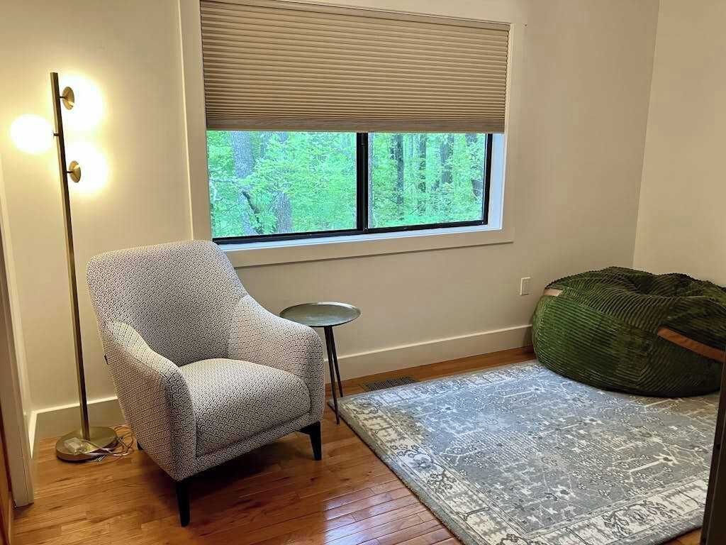 Une pièce comprenant un fauteuil à motifs, une petite table ronde, un pouf vert, un tapis à motifs et une fenêtre avec des stores.