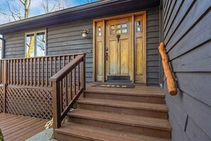 Eine Holztür mit Fußmatte mit der Aufschrift 'HELLO', eine Holztreppe, die zu der Tür hinaufführt, und ein Holengeländer auf der linken Seite.