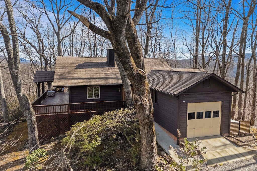 Una baita con veranda, garage e un grande albero in primo piano.
