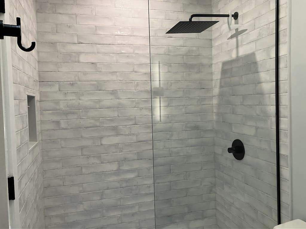 Una ducha con cabezal de ducha negro instalado en la pared y pared cubierta de azulejos blancos.