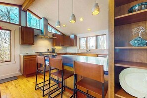 Eine moderne Küche mit Holzausbau, einer zentralen Kochinsel mit Barhocker, Pendelleuchten und großen Fenstern.