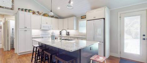 Una cocina con gabinetes blancos, una isla central con taburetes, un refrigerador e iluminación colgante.