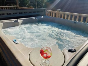 Ein Whirlpool mit einem Glas rotem Getränk und Eiswürfeln, umgeben von einer Holzterrasse und einem Haus im Hintergrund.