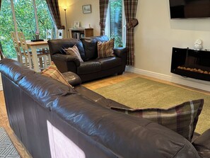 Ein Wohnzimmer mit einem dunkelbraunen Ledersofa, einem Fernseher und Blick auf Grünflächen.