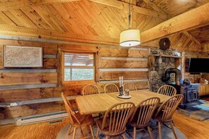 Ein rustikaler Holzhäuschen-Innenraum mit Esstisch, Stühlen, einem Kamin aus Stein und einem Fernseher.