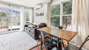 Un espace repas comprenant une table en bois, des chaises noires, un canapé gris avec des coussins et de grandes fenêtres avec des rideaux.