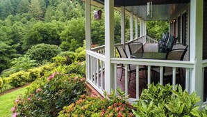 A covered porch with outdoor furniture, surrounded by lush greenery and a view of a forested hillside. - Angel View (Cullowhee)