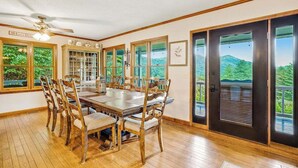 A dining room with a large wooden table and chairs, a ceiling fan, and large windows with a view of greenery. - Angel View (Cullowhee)