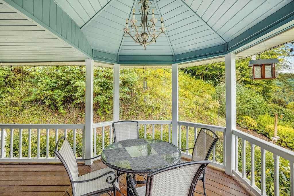 A covered outdoor area with a table and chairs, surrounded by white railings, overlooking a lush green landscape.